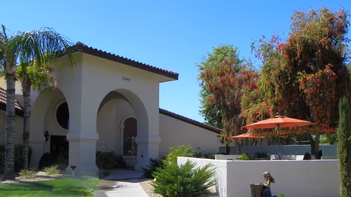 Front entrance of a Mediterranean-style senior living building with arched portico, palm trees, and an outdoor patio with orange umbrellas.