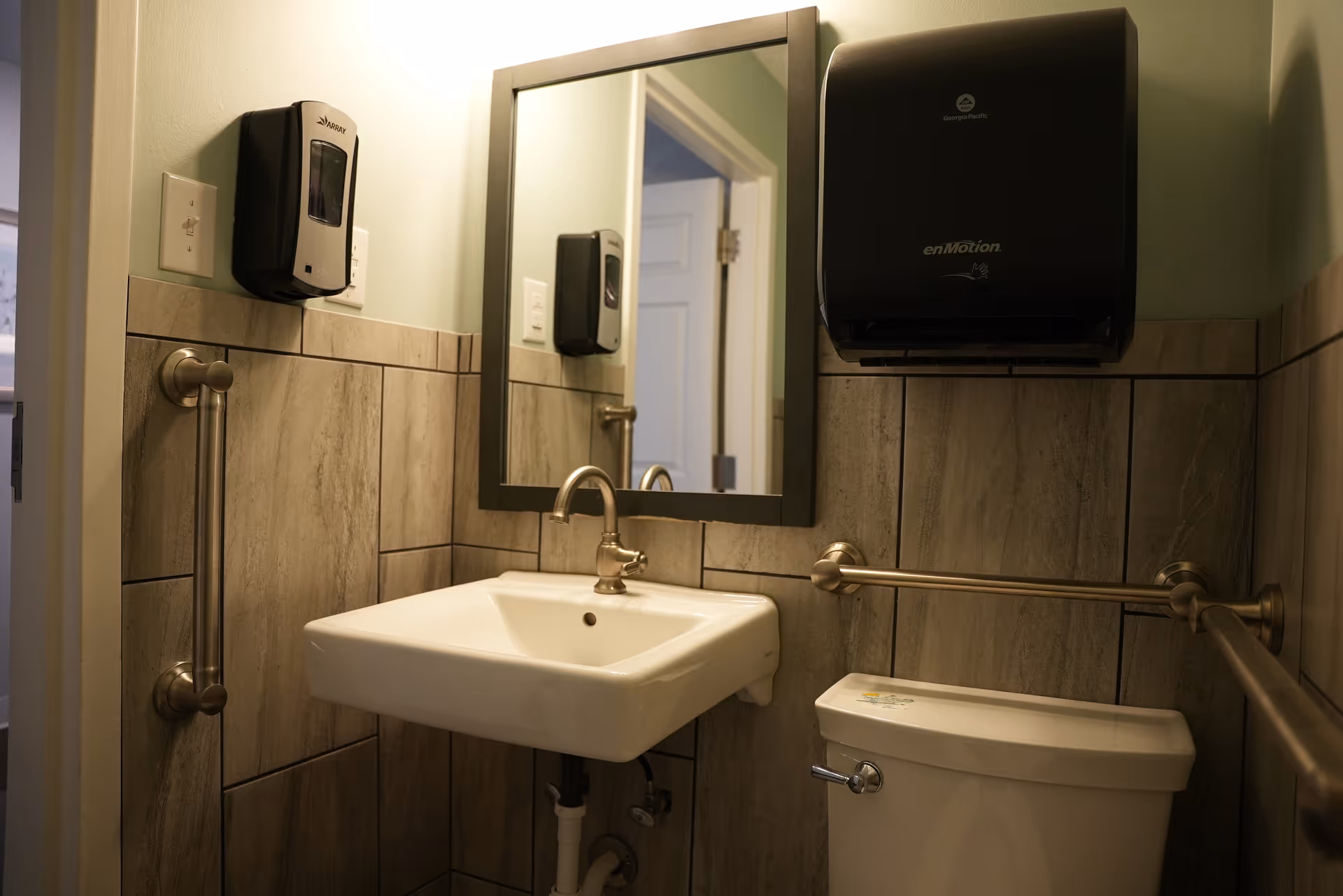 Small bathroom with a white sink, a mirror above it, a toilet, and beige tiled walls. There are metal grab bars on the walls near the sink and toilet, a soap dispenser mounted on the wall, and a black paper towel dispenser above the toilet.