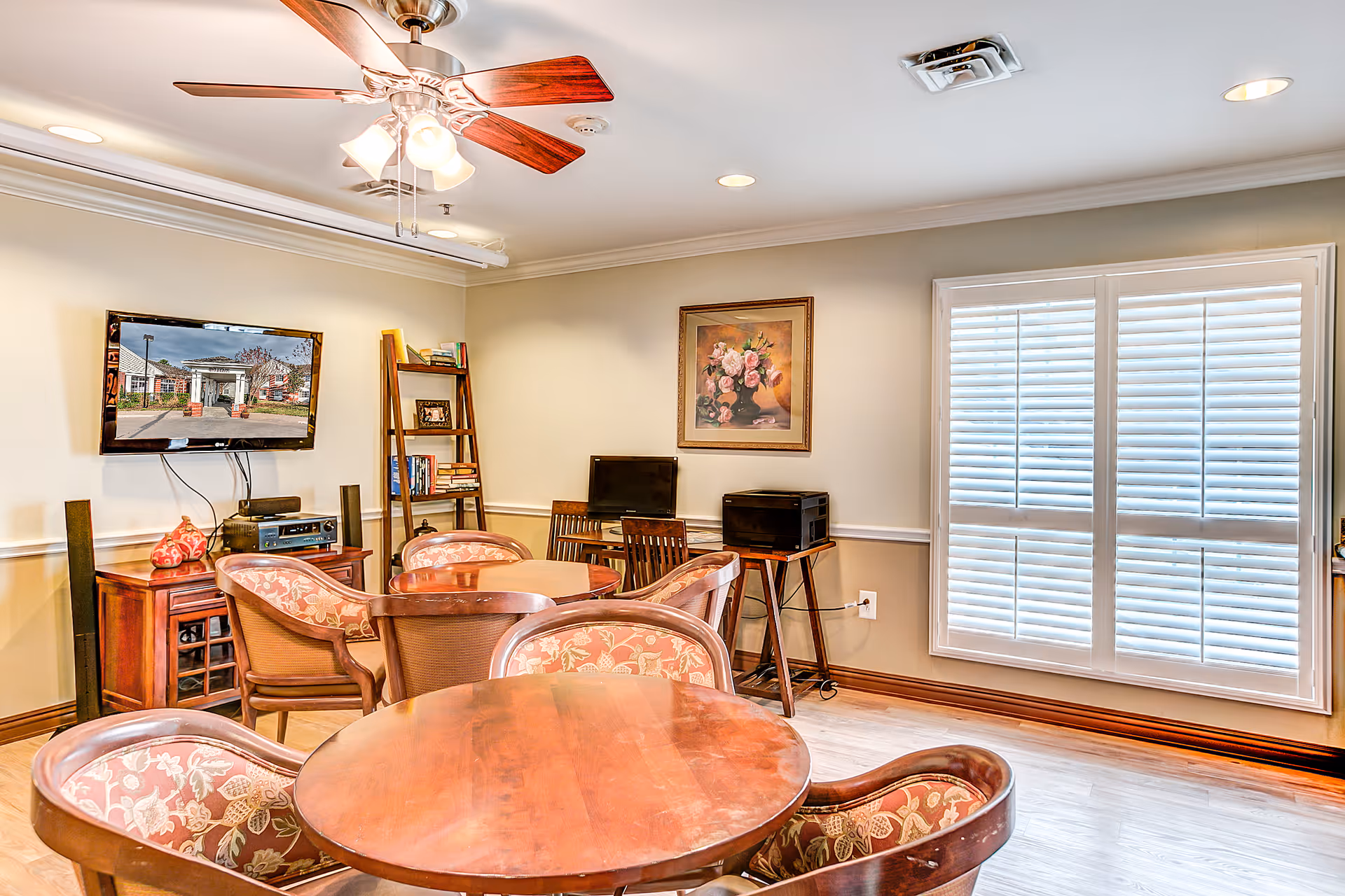 A cozy common area with round wooden tables and floral upholstered chairs. A ceiling fan with lights hangs above. On one wall, there is a flat-screen TV mounted above a wooden cabinet, and a ladder-style bookshelf with books and framed photos. Another wall has a framed painting of flowers and a window with white plantation shutters letting in natural light. The floor is light wood, and the room has a warm, inviting atmosphere.