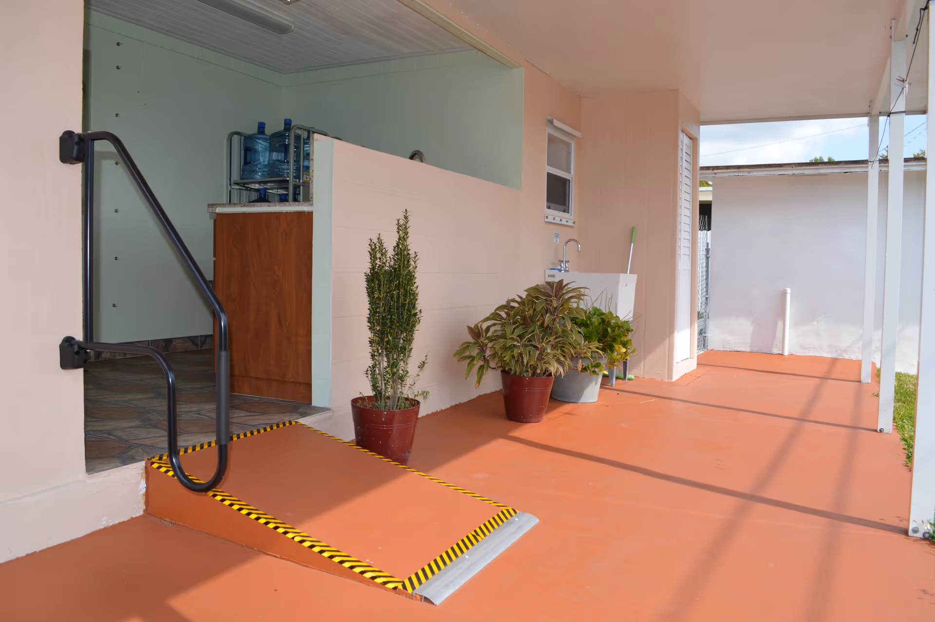Outdoor covered patio area with a small ramp featuring black and yellow caution tape, potted plants, a sink with a faucet, and a mop leaning against the wall. The floor is painted orange and there is a handrail next to the ramp.