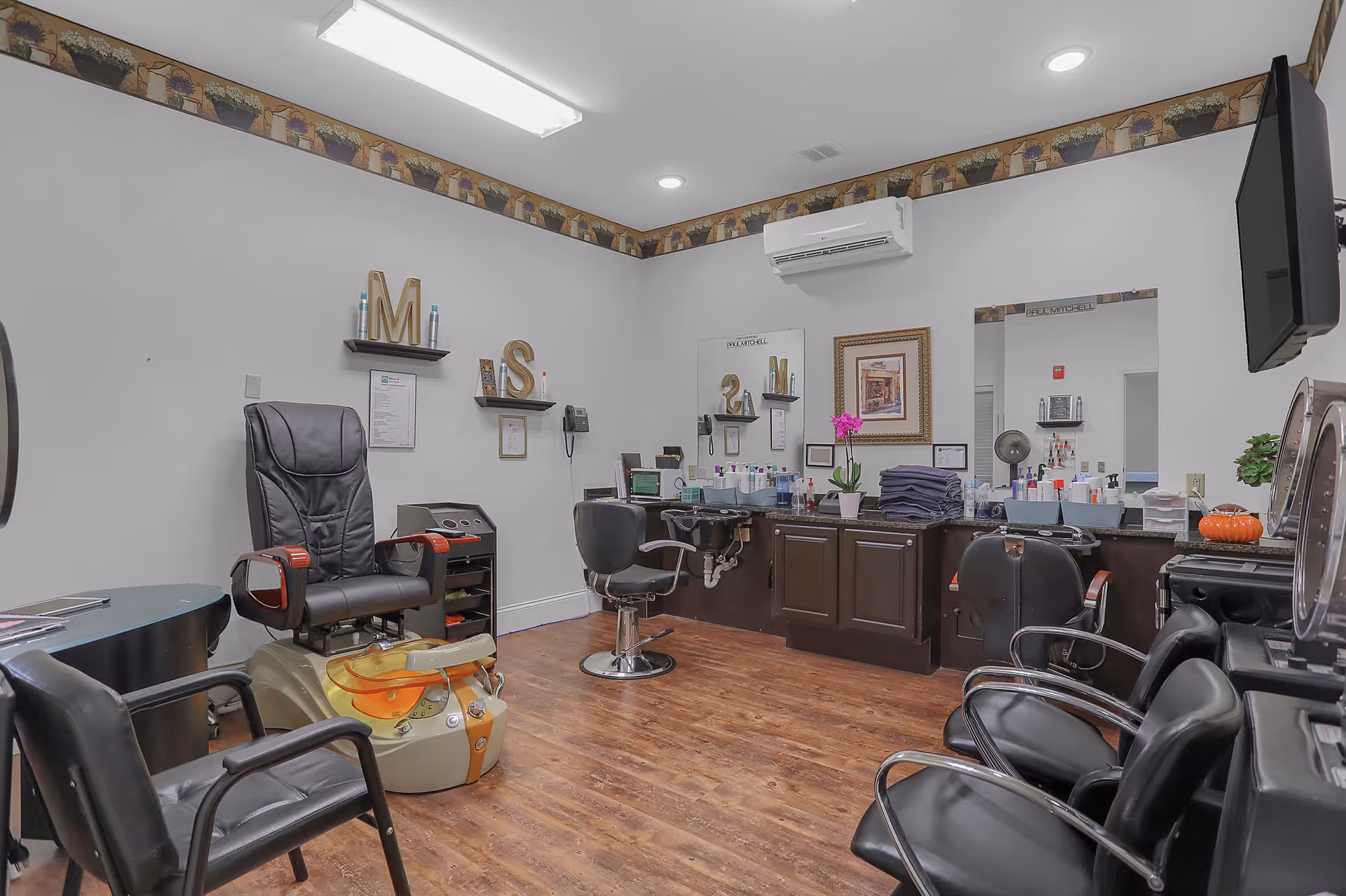 Interior view of a salon room with multiple black salon chairs, a pedicure chair with a foot bath, a large mirror on the wall, shelves with hair products, and a wooden floor. The room has white walls with a decorative border near the ceiling and an air conditioning unit mounted on the wall.