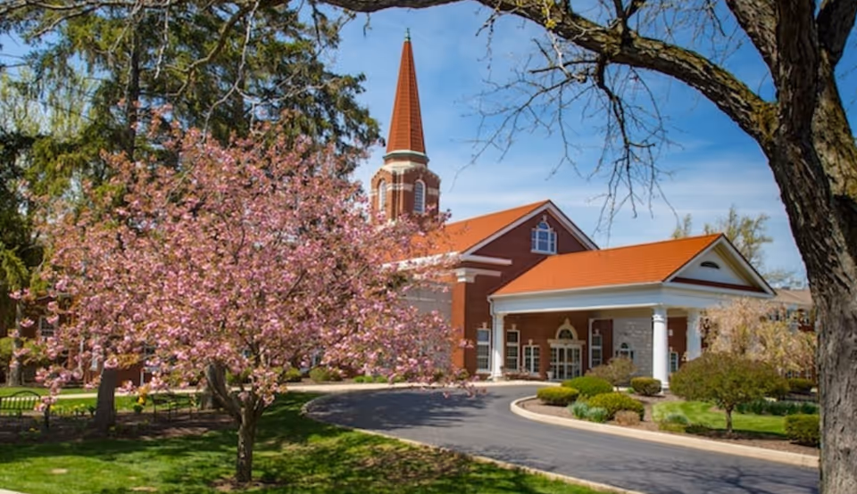 Exterior view of Peabody Retirement Community building with a red roof and white columns at the entrance. A blooming tree with pink flowers is in the foreground, along with other trees and green landscaping under a clear blue sky.