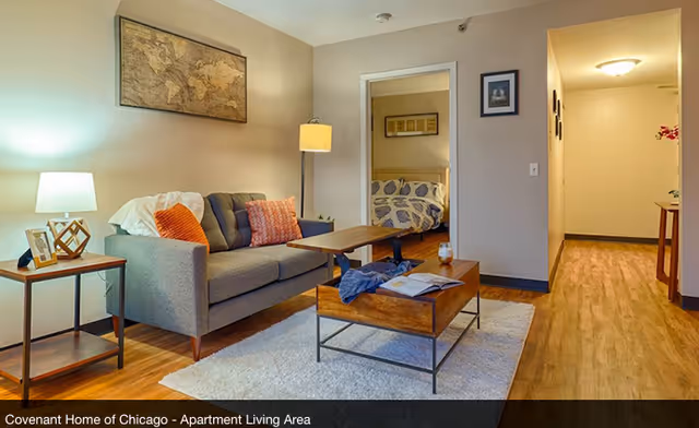 A cozy apartment living area with a gray sofa adorned with orange and white pillows, a wooden coffee table with an open book and a glass, a side table with a lamp and picture frames, a floor lamp, and a framed world map on the wall. In the background, a bedroom with a bed covered in a patterned duvet is visible through an open doorway. The flooring is wood, and there is a hallway leading to other rooms.