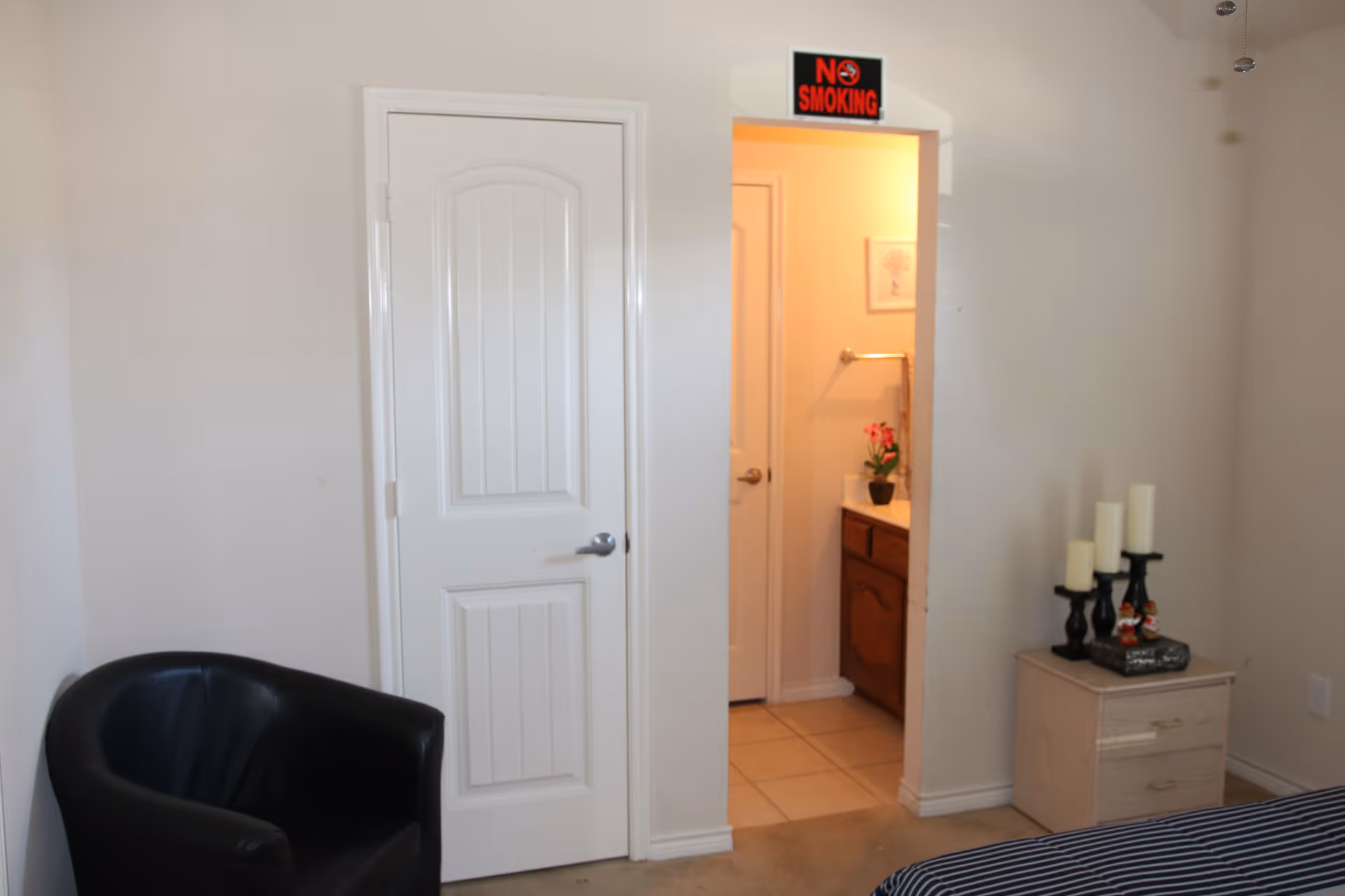 A room with a black leather chair in the corner, a white door, and an open doorway leading to a bathroom with a wooden vanity, a potted plant, and a towel rack. A 'No Smoking' sign is above the bathroom doorway. A small light-colored nightstand with several candles on black candle holders is next to a bed with a striped bedspread.