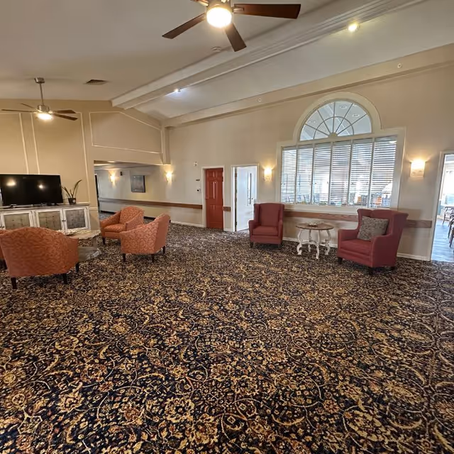 A spacious common area with patterned carpet, several upholstered chairs in shades of red and orange, a small round table, a flat-screen TV on a white cabinet, ceiling fans, and large windows with blinds allowing natural light to enter.