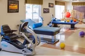Rehabilitation exercise room with a recumbent bike, two treatment tables and colorful therapy balls near windows.