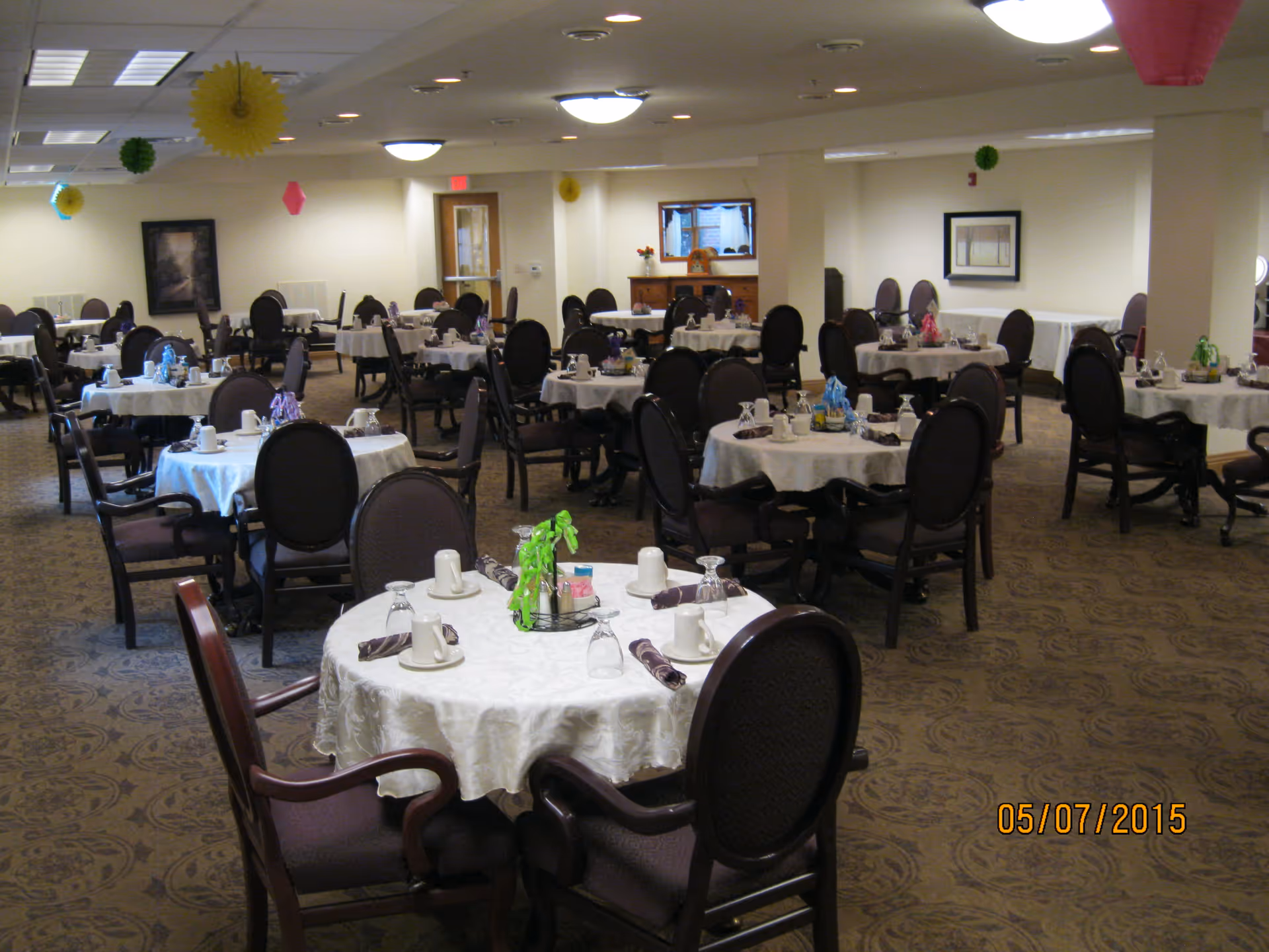 A spacious dining room with multiple round tables covered with white tablecloths, each set with cups, glasses, napkins, and small centerpieces. The room has carpeted floors, beige walls, and ceiling lights. Decorative hanging paper ornaments are visible from the ceiling.