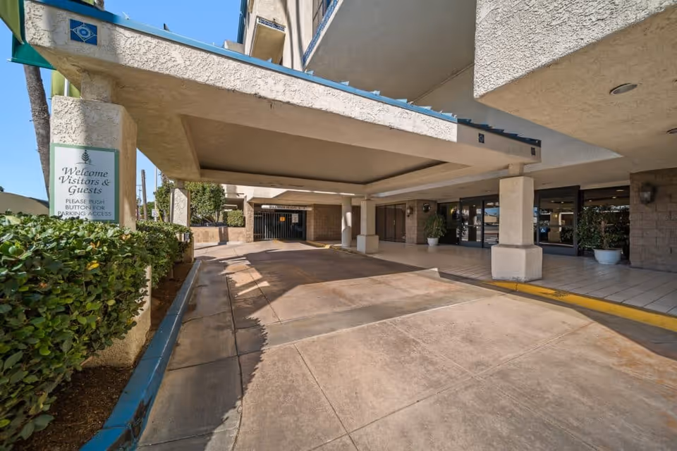 Covered drop-off driveway and entrance with supporting columns, potted plants, and a 'Welcome Visitors & Guests' sign.