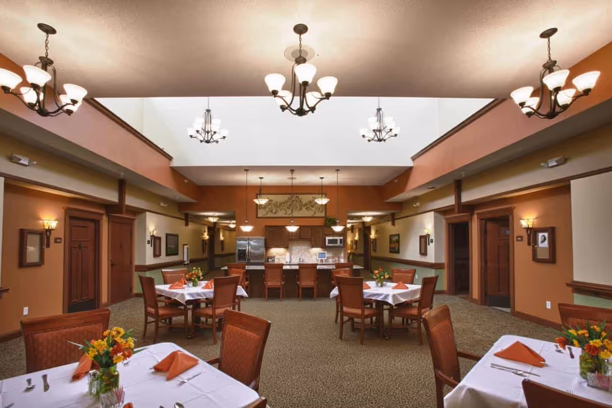 Dining room with tables set with white tablecloths, chairs, floral centerpieces, chandeliers, and a kitchen area in the background.