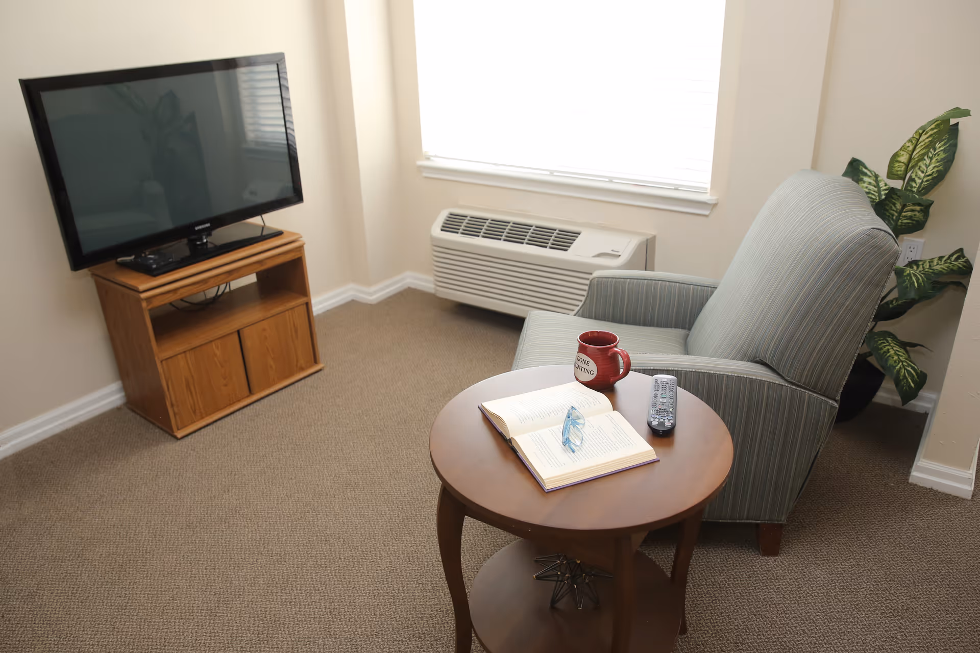 Small sitting area with a striped armchair next to a round side table holding a book, mug and remote, and a TV on a wooden stand by a window.