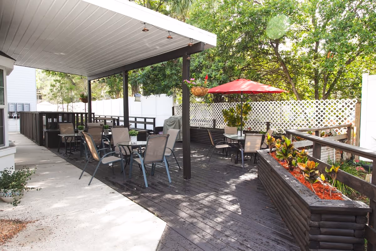Outdoor patio area with several tables and chairs, some under a covered roof and others under a red umbrella. The patio is surrounded by a white lattice fence and greenery, with a planter box containing small plants along the wooden deck.
