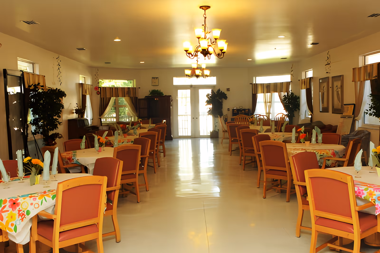 A spacious dining room with multiple tables covered in colorful floral tablecloths and neatly folded napkins. Each table is surrounded by wooden chairs with red cushions. The room is well-lit with ceiling lights and a chandelier in the center. Windows with curtains line the walls, and there are plants and framed pictures decorating the space.