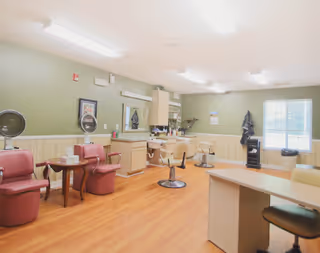 Interior view of a salon area in a senior living facility with several salon chairs, mirrors, and small tables. The room has light green walls with wood paneling, a window letting in natural light, and a wooden floor.