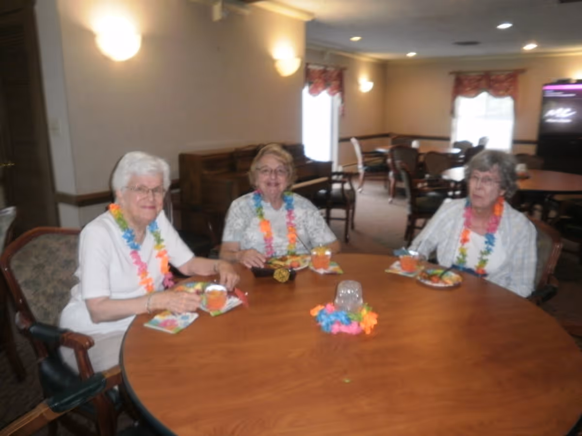 Three elderly women wearing colorful leis are sitting around a round wooden table in a dining area. They have plates of food and drinks in front of them. The room has multiple tables and chairs, with windows covered by curtains in the background.