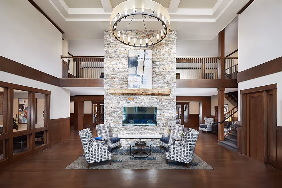 Spacious senior living facility common area with a tall stone fireplace in the center, surrounded by four patterned armchairs and a round coffee table on a rug. The room features high ceilings with a large circular chandelier, wooden paneling, and a second-floor balcony overlooking the space.