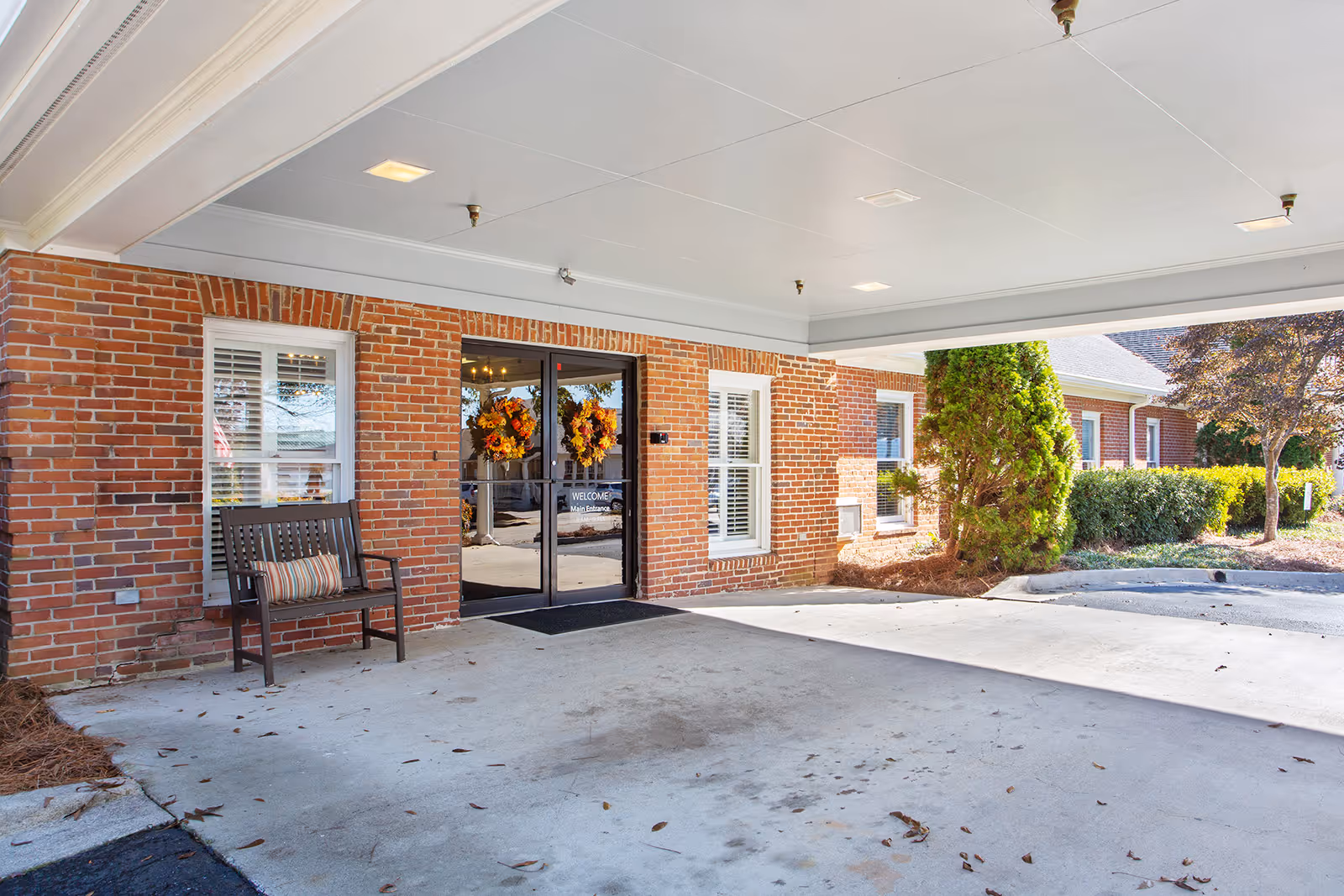 Covered entrance area of a brick building with double glass doors decorated with autumn wreaths. A wooden bench with a striped cushion is placed next to a window with white shutters. There are shrubs and trees in the landscaped area beside the building.