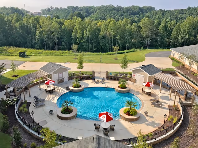 Aerial view of a circular outdoor swimming pool surrounded by a concrete deck with several seating areas, tables with red and white umbrellas, and pergolas. The pool area is enclosed by a black metal fence and landscaped with small trees and plants. Beyond the pool area, there is a grassy field and a dense tree line in the background.