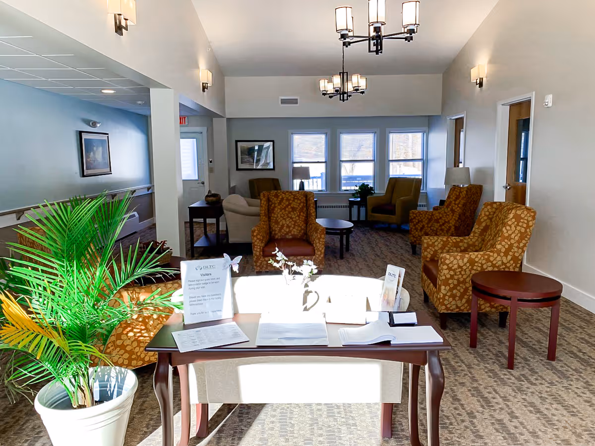 A bright and welcoming common area in a senior living facility with patterned armchairs, a small table with visitor sign-in materials, potted plant, and large windows letting in natural light.