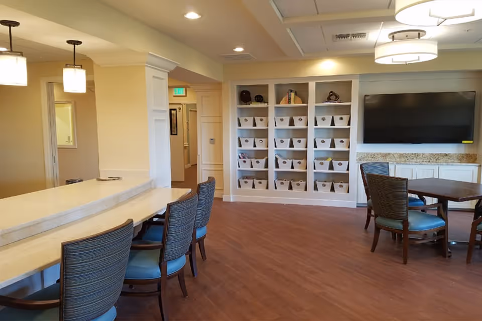 Bright communal room with a long counter and chairs, a wall of cubby shelves, tables, and a mounted TV.