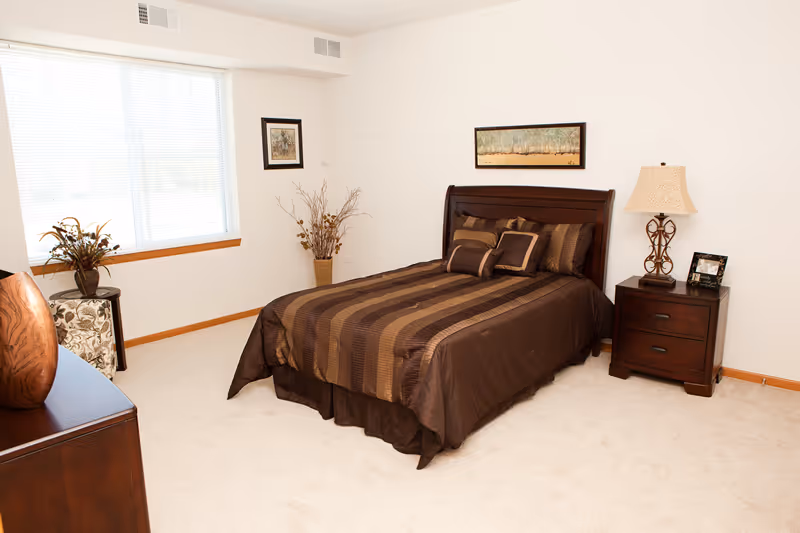 A bedroom with a neatly made bed featuring brown striped bedding and multiple pillows. There is a wooden nightstand with a lamp and a framed photo on it to the right of the bed. A framed landscape painting hangs above the bed. A window with blinds is on the left wall, next to a small table with a floral arrangement. The room has light-colored carpet and white walls.