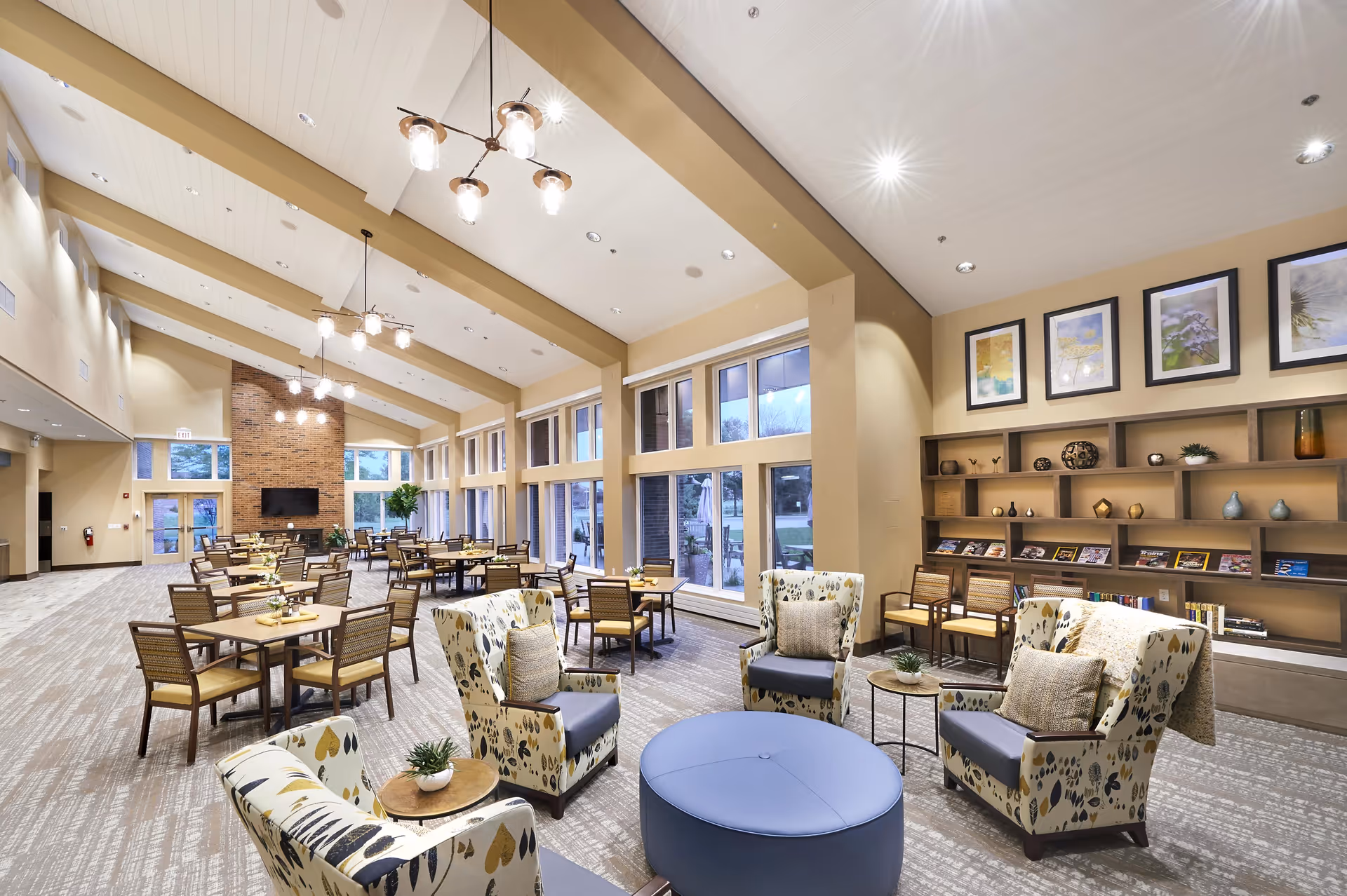 A spacious and well-lit dining and lounge area with multiple tables and chairs arranged neatly. The room features large windows allowing natural light to enter, a brick fireplace with a mounted TV, and modern light fixtures hanging from a high ceiling with exposed beams. There are also comfortable patterned armchairs and a round blue ottoman in the foreground, along with a bookshelf displaying decorative items and books.