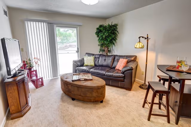A cozy living room with a dark brown leather sofa adorned with two decorative pillows, one green and one orange. In front of the sofa is a brown oval ottoman with a tray on top. To the right, there is a tall floor lamp with a stained glass shade and a small dining area with a dark wooden table and two stools. On the left side, a flat-screen TV sits on a wooden stand next to a small red shelf with plants. A sliding glass door with vertical blinds lets in natural light and shows greenery outside.