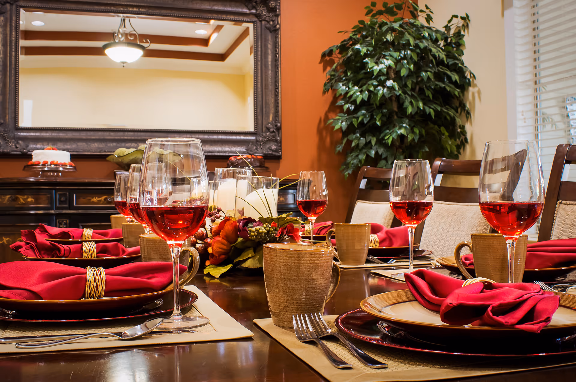 A dining table set for a meal with red wine glasses, beige mugs, plates with red napkins, and silverware. The table has a centerpiece with candles and flowers. In the background, there is a large ornate mirror, a sideboard with cakes, and a large green plant next to a window with blinds.