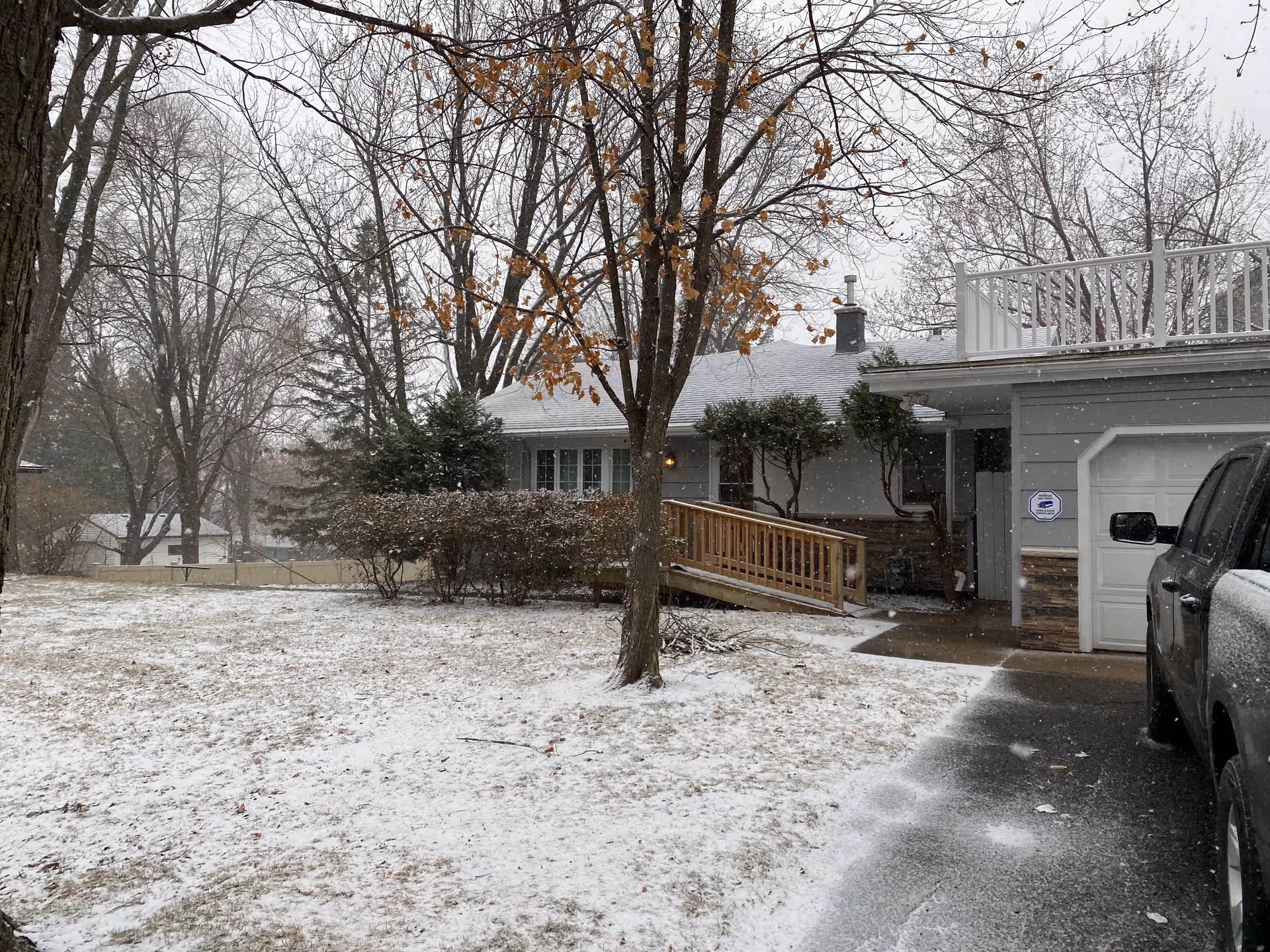 Snow-dusted front yard and driveway leading to a single-story house with a wooden accessibility ramp and attached garage during light snowfall.