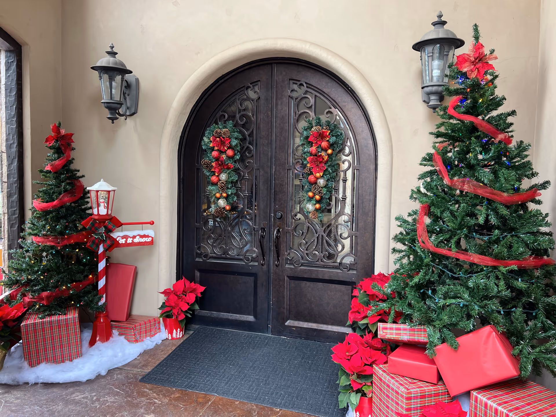 Arched double entry doors decorated with Christmas wreaths, flanked by two decorated Christmas trees, wrapped gifts and poinsettias.