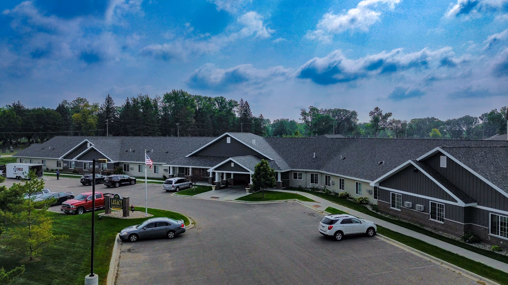 Exterior view of The Legacy Assisted Living facility in Morris, MN, showing a single-story building with a large parking lot in front. Several cars are parked, and there is a flagpole with an American flag near the entrance. Trees and greenery surround the building under a partly cloudy sky.