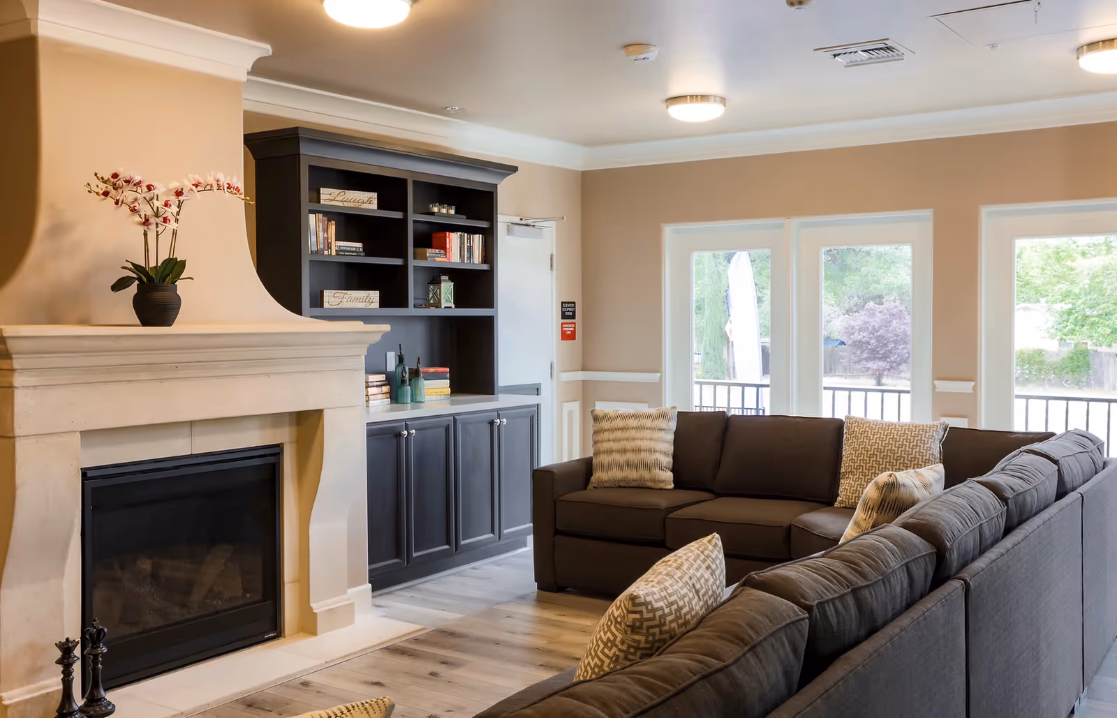 A cozy living room with a large dark gray sectional sofa adorned with patterned pillows, a beige stone fireplace with a potted orchid on the mantel, and a dark wood built-in bookshelf filled with books and decorative items. Large windows in the background let in natural light and show greenery outside.