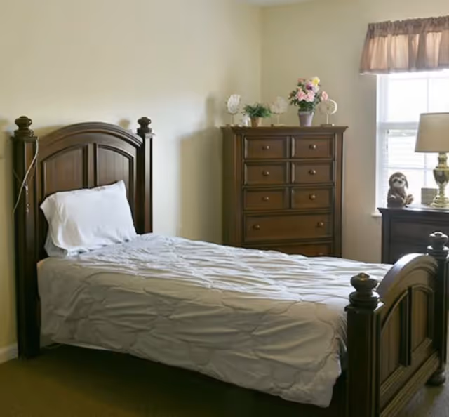A neatly made single bed with a white pillow and quilt in a room with beige walls. Next to the bed is a wooden dresser with six drawers, decorated with a potted plant and flowers. A window with a brown valance lets in natural light, and a nightstand with a lamp and a small stuffed animal is beside the window.
