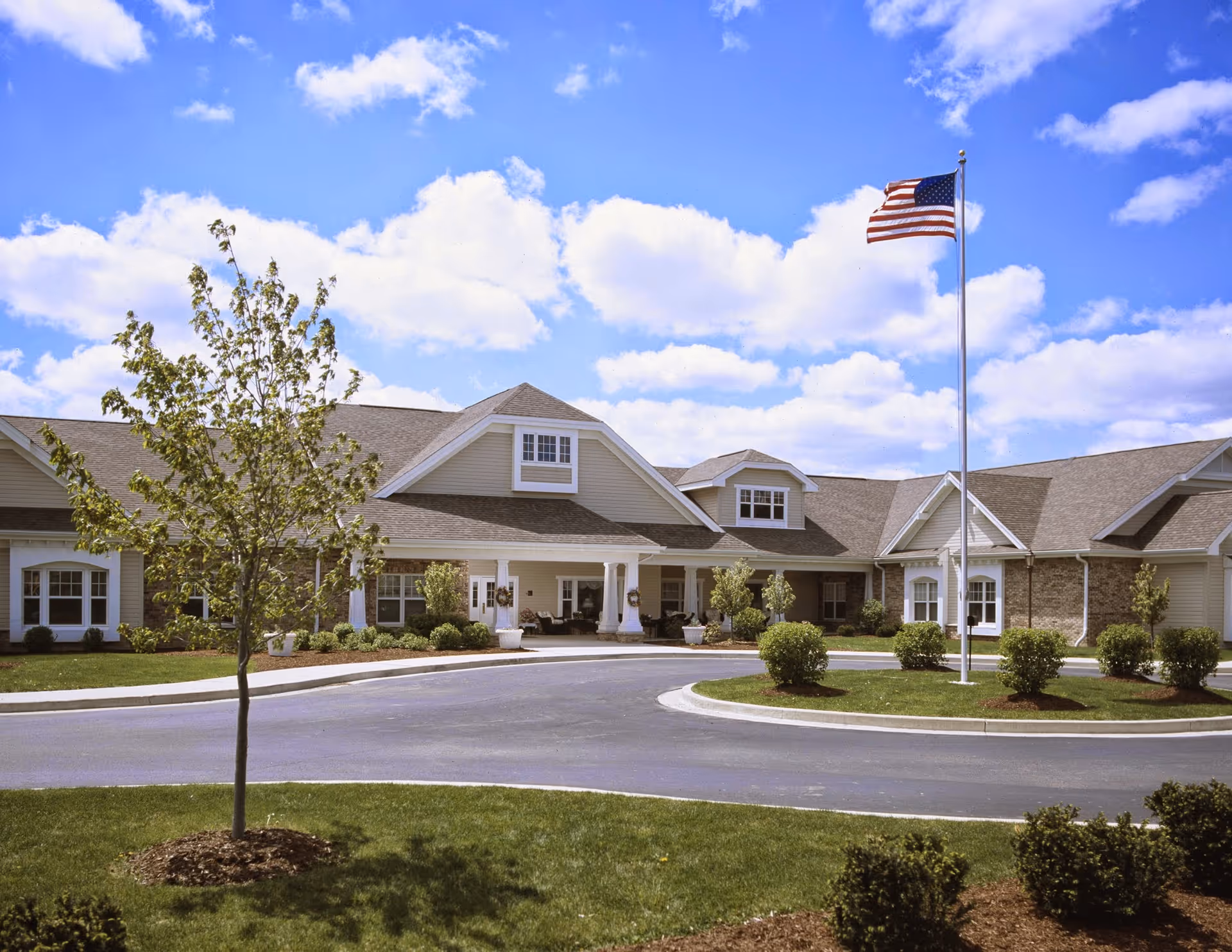 Exterior view of a single-story senior living facility building with beige siding and brick accents, a circular driveway, landscaped bushes and trees, and an American flag on a flagpole under a partly cloudy blue sky.