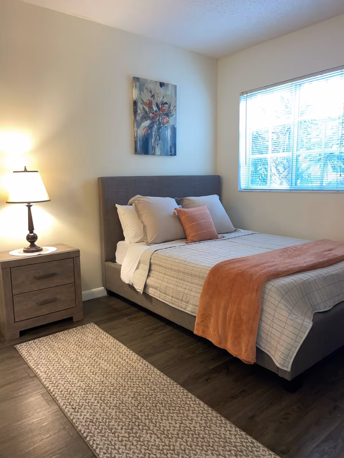 A neatly made bed with a gray upholstered headboard, beige and white pillows, and an orange throw blanket in a bedroom with a wooden nightstand and a lamp. A painting of flowers hangs on the wall above the bed, and a window with blinds lets in natural light.