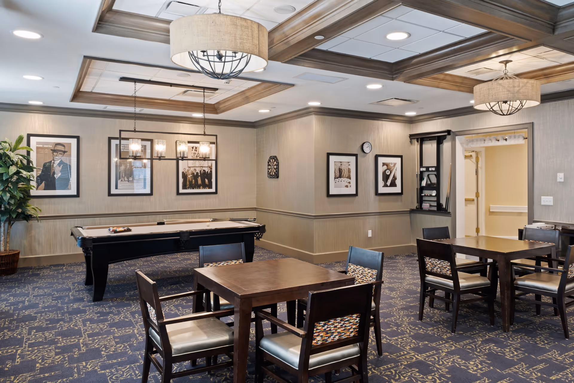 A well-lit recreational room featuring a pool table with billiard balls arranged on it, several wooden tables with chairs, framed black and white photographs on the walls, a dartboard, and decorative ceiling lights. The room has patterned carpet flooring and neutral-colored walls with wooden trim.