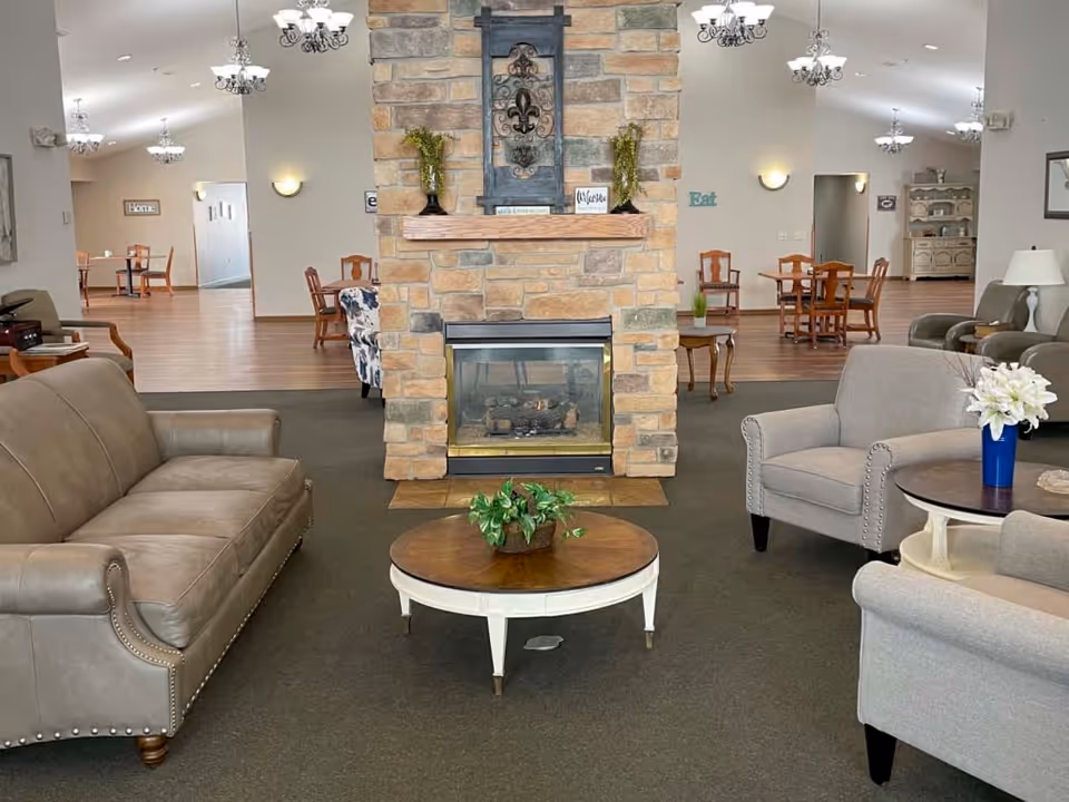 A cozy senior living facility common area with a stone fireplace in the center. Surrounding the fireplace are comfortable seating options including a gray leather sofa, two gray armchairs, and a round wooden coffee table with a small green plant. In the background, there are wooden dining tables and chairs on a wood floor, with soft lighting from chandeliers and wall sconces. The space is decorated with small plants and welcoming signs.