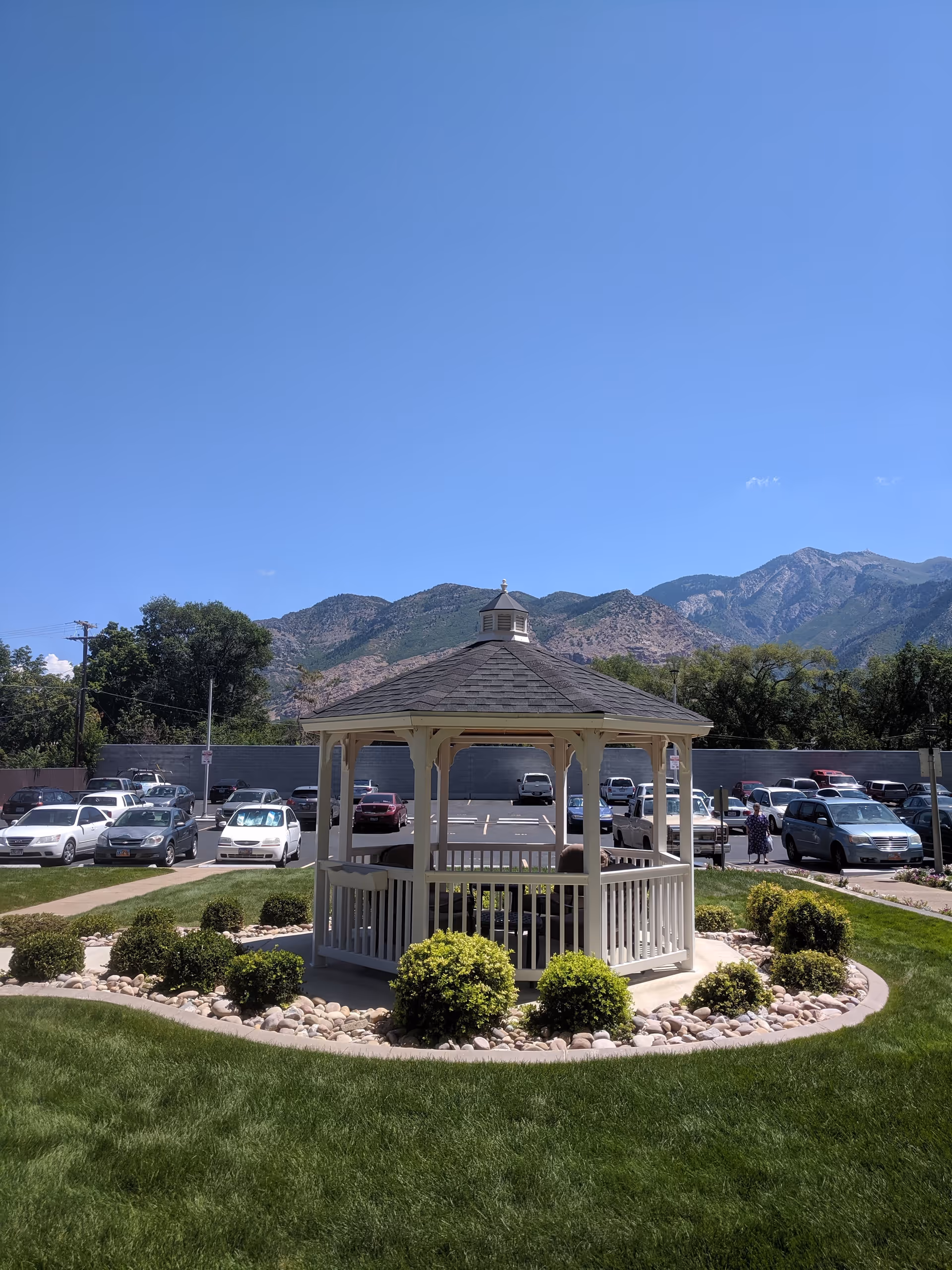 A white gazebo surrounded by neatly trimmed bushes and rocks in a circular garden area with green grass. Behind the gazebo is a parking lot with several parked cars and a few people walking. In the background, there are tall mountains under a clear blue sky.