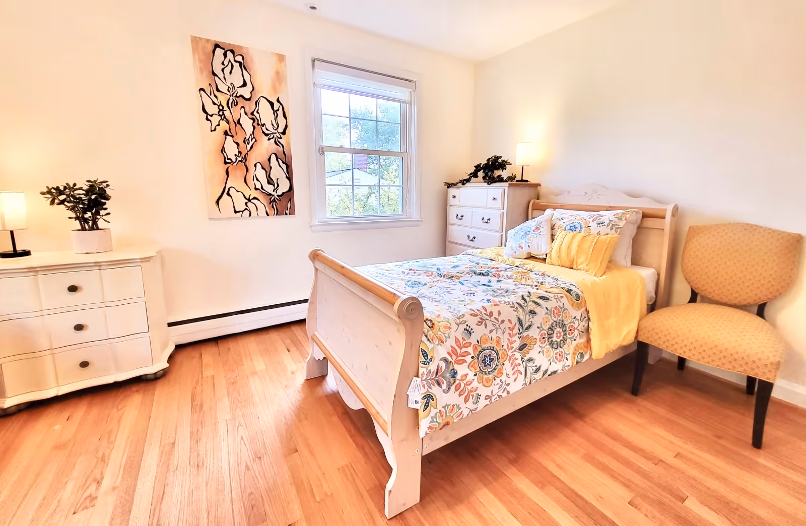 A bright and cozy bedroom with a wooden bed featuring floral bedding and yellow accents. The room has hardwood floors, a window letting in natural light, a white dresser with a lamp and plant, a chest of drawers with a lamp and greenery, a patterned chair, and a floral painting on the wall.