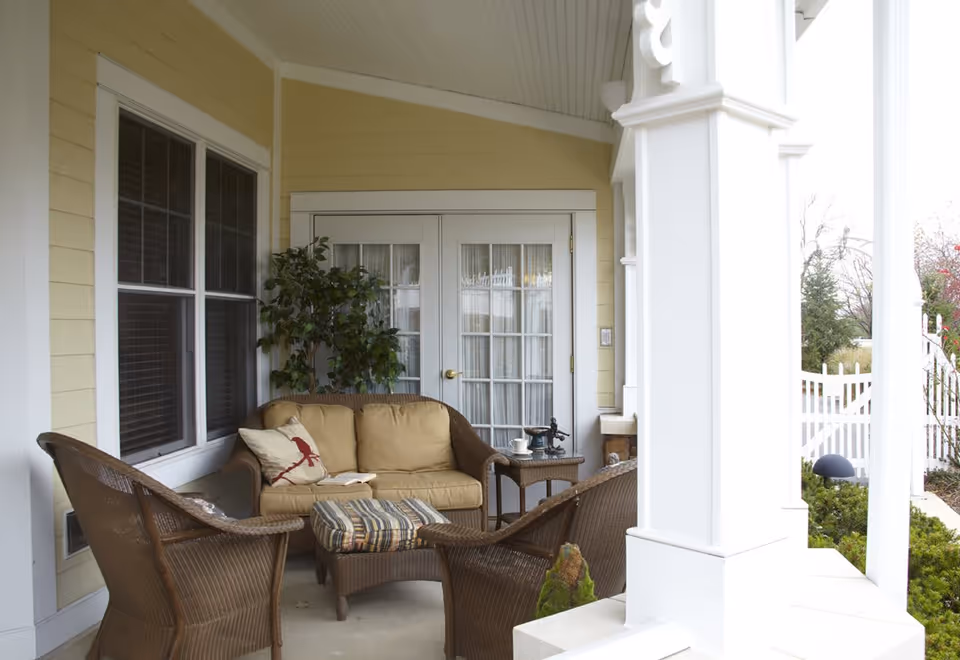 A cozy outdoor porch area with wicker furniture including a cushioned loveseat, two chairs, and a striped ottoman. There is a small side table with a decorative item and a cup, a potted plant in the corner, and a white picket fence visible in the background.