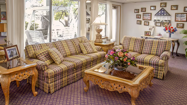 A cozy living room with two plaid sofas facing each other, a wooden coffee table with a floral arrangement in the center, a side table with a lamp, and a wall decorated with framed photos and a folded American flag display case. Large windows and a glass door provide a view of an outdoor swing and greenery.