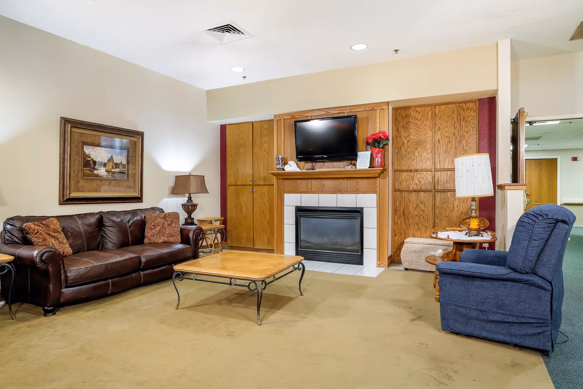 A cozy senior living facility common area with a brown leather sofa, a blue upholstered armchair, a wooden coffee table, and a fireplace with a mounted flat-screen TV above it. There are wooden cabinets on either side of the fireplace, a table lamp on a side table, and a framed painting on the wall.