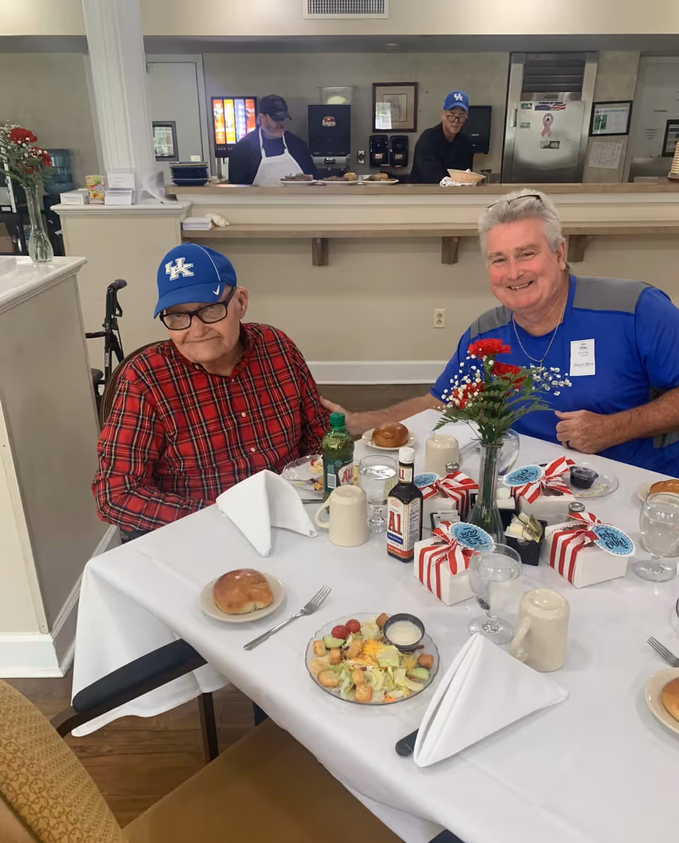 Two elderly men sitting at a dining table in a senior living facility. One man is wearing a red plaid shirt and a blue cap, while the other is in a blue shirt with a name tag. The table is set with plates of salad, rolls, drinks, condiments, and small gift boxes with red and white ribbons. In the background, two staff members are behind a counter preparing or serving food.