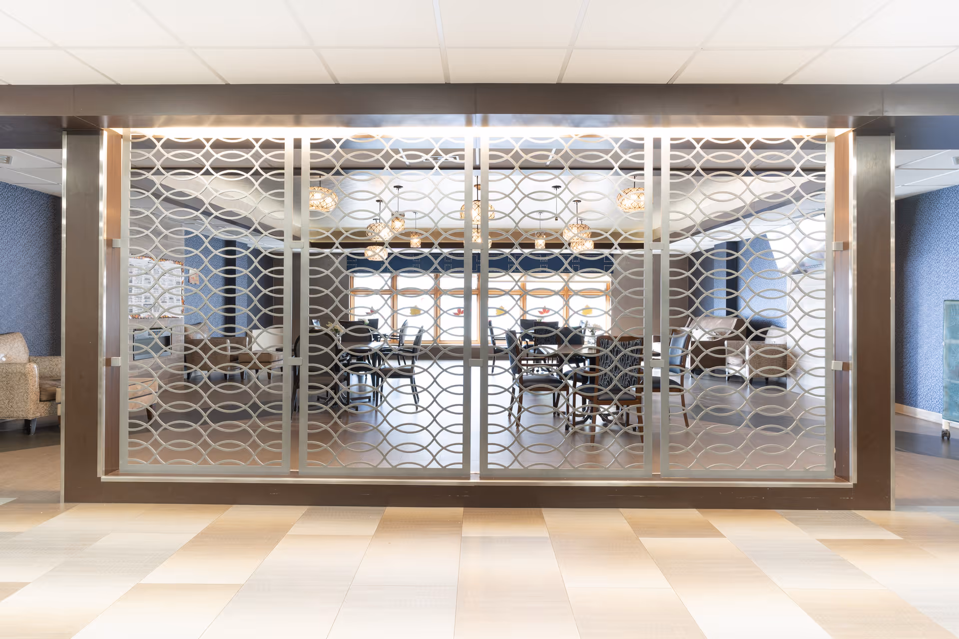 View through a decorative metal screen into a well-lit common area with multiple tables and chairs, pendant lights hanging from the ceiling, and large windows letting in natural light. The room has blue walls and comfortable seating along the sides.