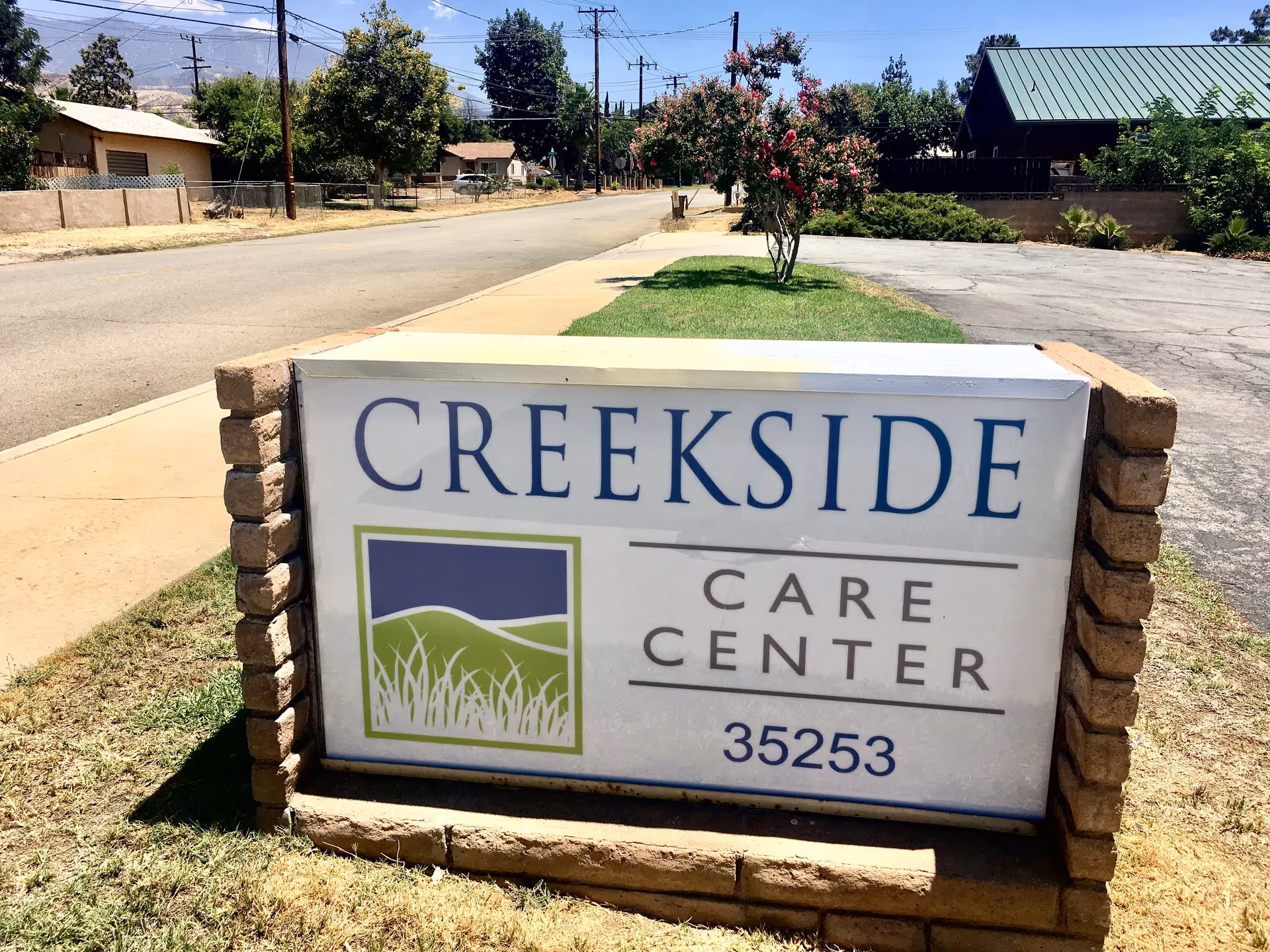 A roadside sign that reads "Creekside Care Center 35253" mounted on a brick base with a neighborhood street and trees behind it.