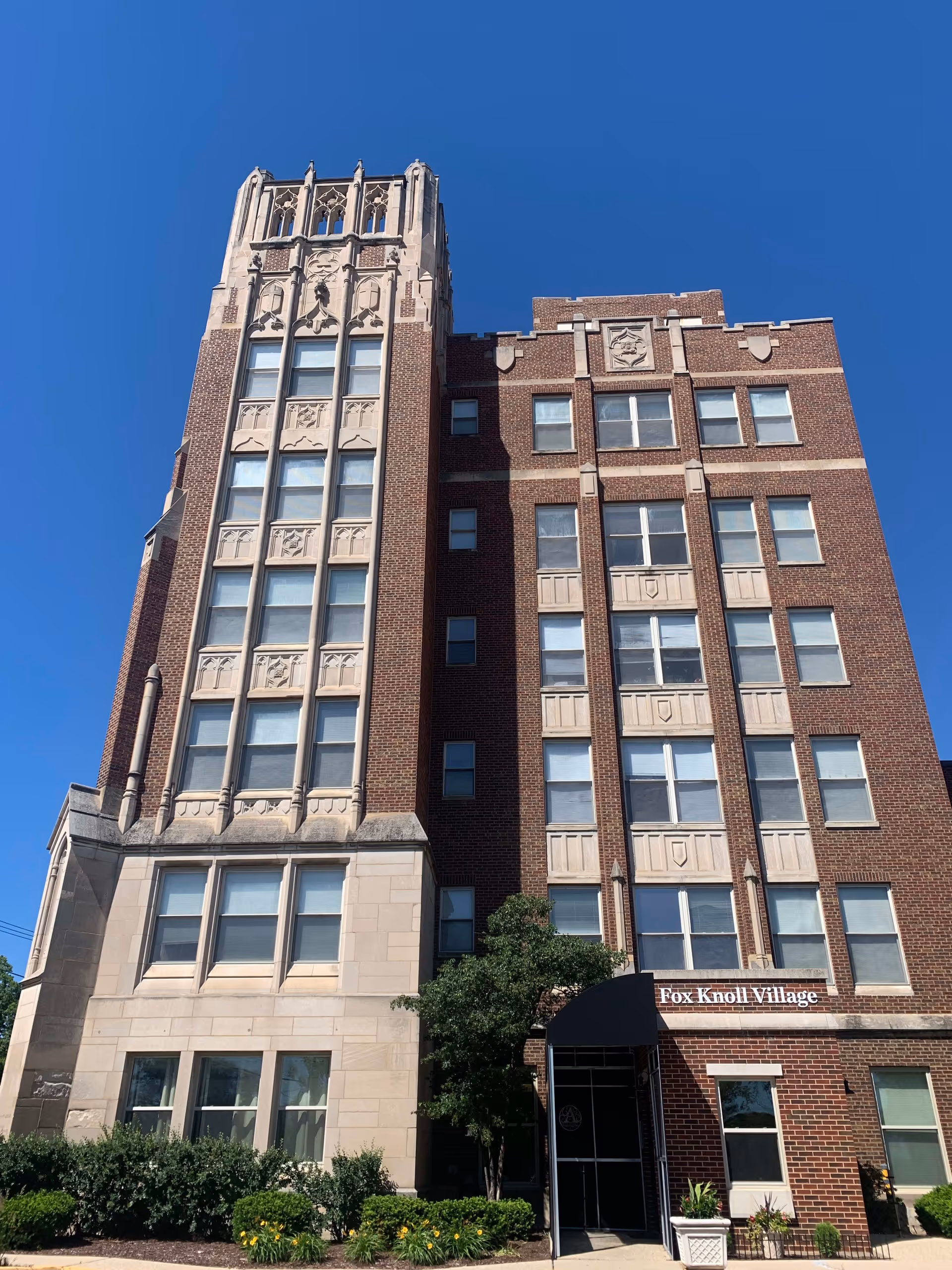 Exterior view of a multi-story brick building with decorative stone accents under a clear blue sky. The building has many windows and a black awning above the entrance with the sign 'Fox Knoll Village'. There are shrubs and flowers planted in front of the building.