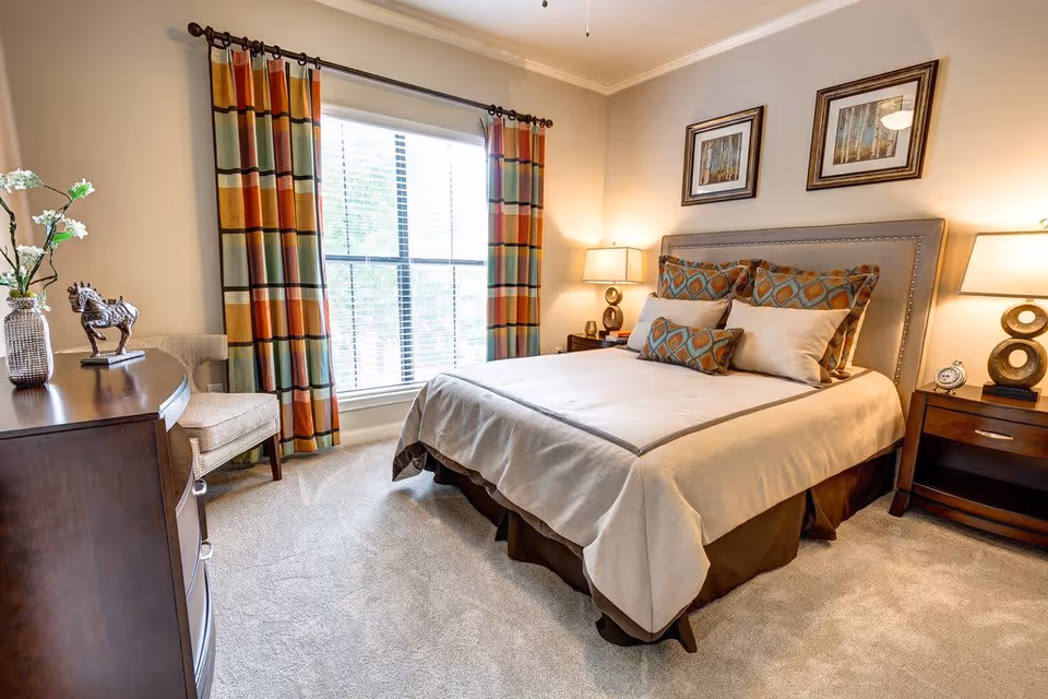 A cozy bedroom with a neatly made bed featuring patterned pillows and a beige bedspread. The room has a large window with colorful striped curtains, two wooden nightstands with lamps, framed artwork on the wall above the bed, a wooden dresser with decorative items, and a beige upholstered chair in the corner.