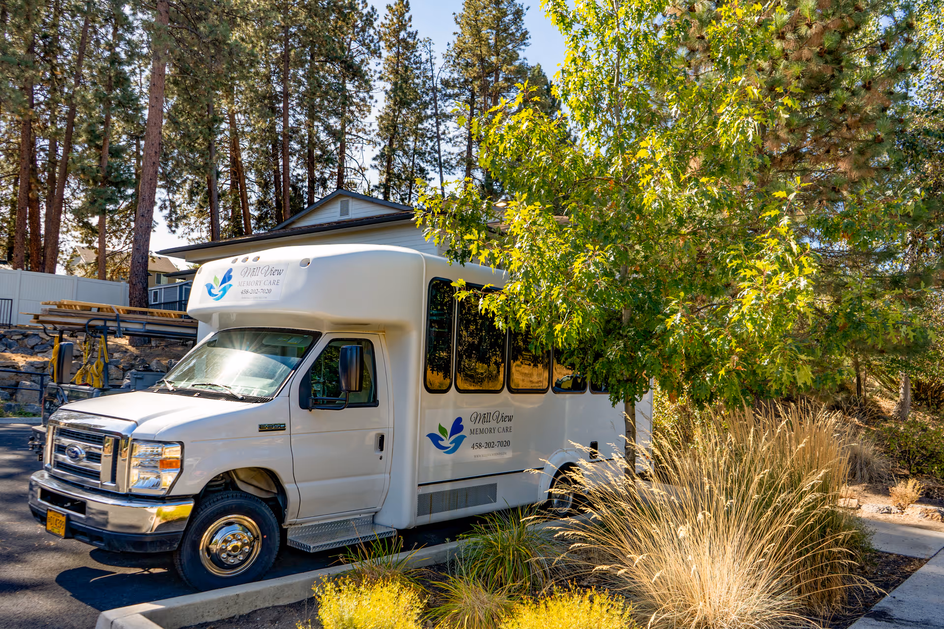 A white shuttle bus parked in a lot surrounded by trees and landscaping. The bus has the logo and text 'Mill View Memory Care' on its side. The area is sunny with shadows cast by the trees.