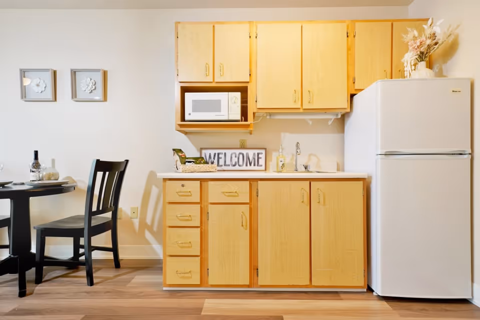 A small kitchen area with light wood cabinets, a white microwave, a white refrigerator, and a countertop with a sink. A decorative 'WELCOME' sign and a basket are on the counter. To the left, there is a black dining table with two black chairs, set with plates, wine glasses, and a bottle of wine. Two framed floral artworks hang on the wall above the dining table.