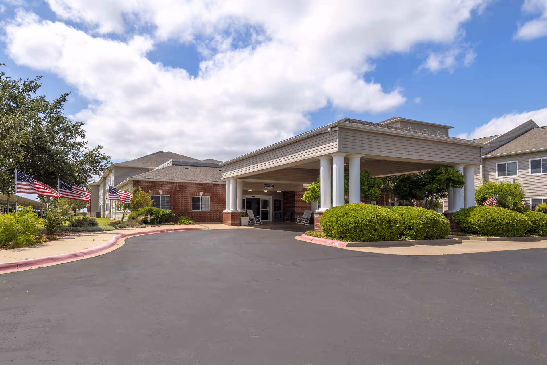 Front entrance of the Lyndale San Angelo Senior Living building with a covered porte-cochere, white columns, landscaping and American flags.