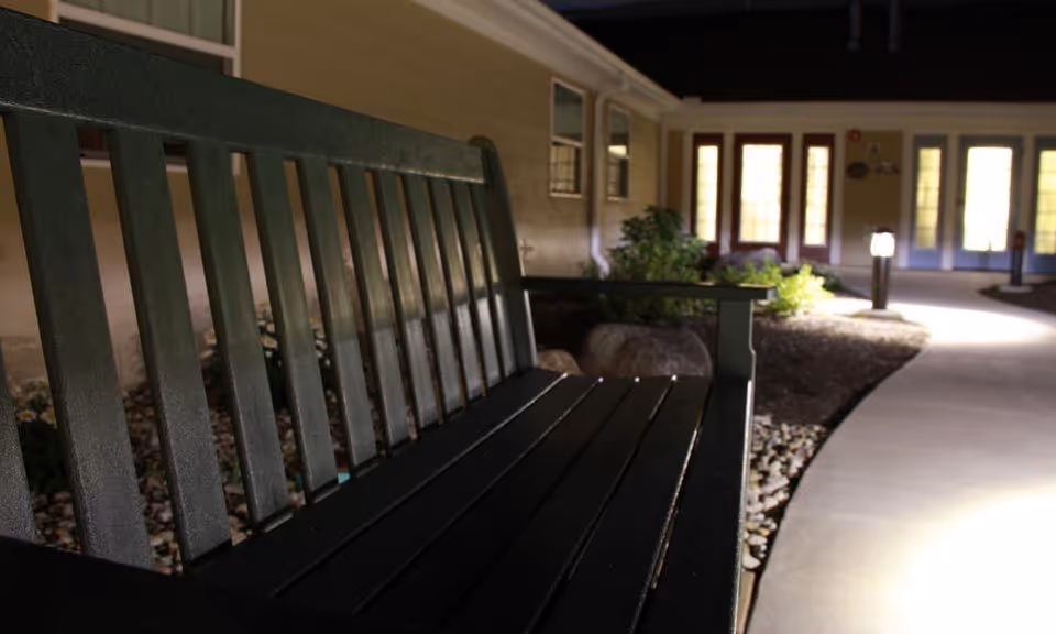 A close-up view of a dark green outdoor bench along a curved concrete pathway at night, with illuminated garden lights and the exterior of a building with multiple windows and doors in the background.
